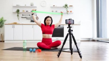 Kırmızı spor elbiseli bir kadın ev mutfağındaki çevrimiçi fitness sınıfı için direnç bandı egzersizi yapıyor. Yoga minderinde su şişesi, dambıl, akıllı telefon tripodu ve dizüstü bilgisayarla oturuyor..
