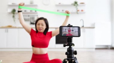 Kırmızı spor elbiseli bir kadın modern mutfakta online fitness kursu sırasında direniş bandı egzersizleri yapıyor. Akıllı telefon ve tripod kurulumu ile fitness videosu çekiliyor.
