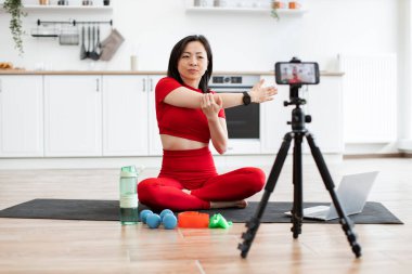 Kırmızı spor elbiseli bir kadın, spor salonunda esniyor. Yoga minderi, egzersiz malzemeleri, su şişesi ve sanal fitness sınıfı için kurulmuş kamera..