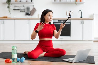 Kadın telefonda çalışmak için çevrimiçi blog kaydediyor. Mutfakta yoga minderinde oturmak, kırmızı antreman kıyafetleri giymek, dizüstü bilgisayar ve tripoyu film çekmek için kullanmak. Çevrimiçi fitness eğitimi ve vloglama kavramı.