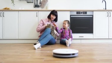Anne ve kızı mutfak zemininde oturmuş robot süpürge çalışırken evcil kediyle oynuyorlar. Parlak ve rahat mutfak dekorasyonu, aile bağlarını geliştirme, evcil hayvanları sevme.
