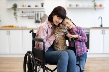 Tekerlekli sandalyedeki gülümseyen anne, sevgi dolu kızını kucaklayan kediyi kucağına alıp evde sıcaklık, bakım ve kapsayıcılığı taşıyor.