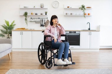 Tekerlekli sandalyedeki genç kadın modern mutfakta oturuyor, düşünceli görünüyor. Dekor, bitki ve modern aletlerle rahat ve şık bir iç mekan..