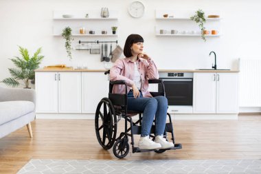 Tekerlekli sandalyedeki kadın, modern mutfak alanında oturuyor, günlük kıyafetler giyiyor, düşünceli görünüyor. Parlak, iyi dekore edilmiş, bitkiler ve mutfak gereçleri ile donatılmış..