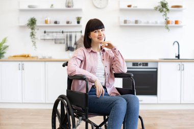 Tekerlekli sandalyede oturan genç bir kadın modern mutfak ortamında sıcak bir şekilde gülümsüyor. Kapsayıcılık, bağımsızlık ve pozitif yaşam kavramı.