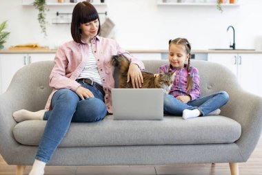 Anne ve kızı dizüstü bilgisayarı ve evcil kedisiyle gri koltukta oturuyor. Modern oturma odasında rahat bir dekorla aile, rahatlama ve bağlanma anlarını tasvir eden sade bir sahne..