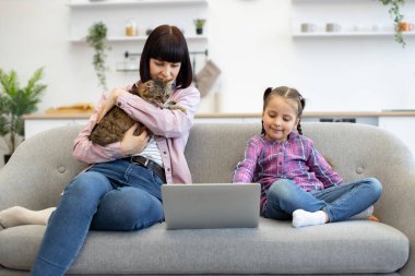 Anne kediyi kucaklarken kız kanepede dizüstü bilgisayar kullanıyor. Rahat atmosferli bir ev. Aile bağları sevgi, teknoloji ve evcil hayvanları gösteriyor..