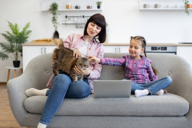 Anne ve kızı dizüstü bilgisayarlarıyla kanepede oturmuş, tüylü kedilerle kaynaşıyorlar. Konforlu ev ortamında rahatlamak. Modern yaşam alanında bitkilerle birlikte kaliteli zaman geçirmekten hoşlanırlar..