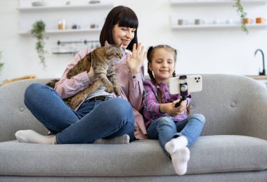 Anne ve kızı kanepede oturup kediyle selfie çekiyorlar. Anne ve kedi kucağında otururken çocuk selfie çubuğunda telefon tutuyor. Rafları ve bitkileri arka planda olan rahat bir ev ortamı