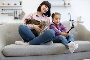 Selfie çubuğu tutan çocuk, sarılan kedi annesi. Anne ve kızı kanepede oturup akıllı telefonla selfie çekiyorlar. Aile bağlarını güçlendirme anlarıyla iç içe yaşıyorlar..