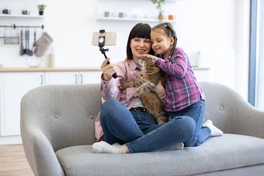 Anne ve kızı kanepede oturup kedi beslerken akıllı telefonuyla selfie çekiyorlar. Ev içinde mutlu aile anları dilerim. Gülümseyen yüzler neşe ve beraberliği ifade eder.