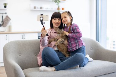 Aile bağı, mutluluk ve evcil hayvan sevgisi anı yakalar. Anne ve kız kanepede kediyle selfie çekerken gülümsüyor. Modern yaşam tarzı, teknoloji ve birliktelik .