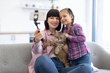 Parlak iç mekan, sıcak ve mutlu bir atmosfer yaratır. Anne ve kızı evde kedileriyle selfie çekerken gülümsüyorlar. Kanepede oturuyorlar ve ailecek vakit geçiriyorlar..
