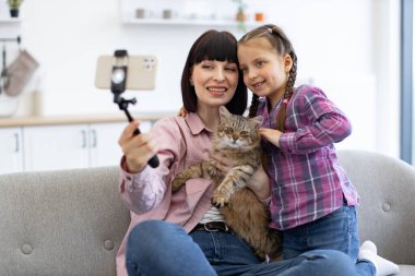 Gülümseyen anne ve kızı kedileriyle selfie çekiyorlar. Kapalı alanda akıllı telefon kullanıyorlar. Aile bağı, evcil hayvan sevgisi ve rahat ev ortamında tasvir edilen modern teknoloji..