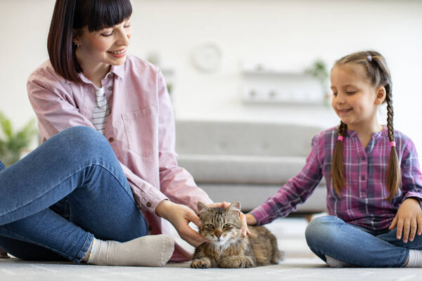 Мать и дочь наслаждаются временем вместе, лаская кошку дома. Оба сидели на полу с яркими улыбками, запечатлевая любовь и радость в приятной гостиной.