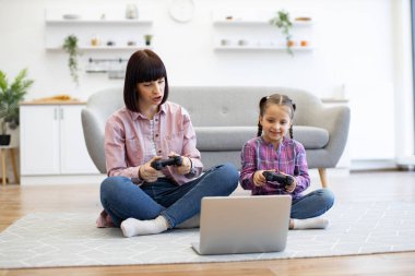 Anne ve kızı oyun kontrolörlerini kullanarak yerde oturup oyun oynuyorlar. Arka planda kanepe ve bitkilerle rahat bir ev, ikisi de kaliteli zaman geçirmekten hoşlanıyor..