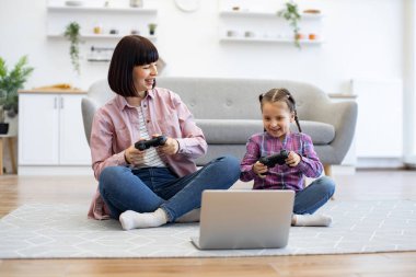 Samimi ve modern ev ortamında oyun kontrolörleriyle gülüp eğleniyorlar. Anne ve kızı birlikte oturma odasında video oyunu oynuyorlar..