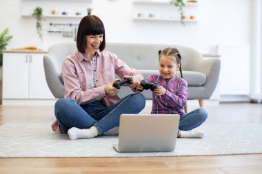 Gülümseyen aile yerde oturup eğleniyor. Anne ve kızı evde bilgisayar oyunu oynayarak eğleniyorlar. Hafif dekorlu ve mutlu ifadeli rahat oturma odası sahnesi.