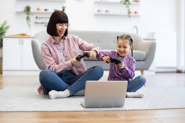 Anne ve kızı oyun kontrolörlerini kullanarak yerde oturup oyun oynuyorlar. Hem gülümseyen hem de eğlenceli aktiviteler yapan samimi oturma odası ortamında.