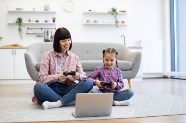 Anne ve kızı, sıcak oturma odasında birlikte video oyunu oynayarak eğleniyorlar. Dizüstü bilgisayar ve kontrolörlerle eğlenirken gülümseyen aile bağı.