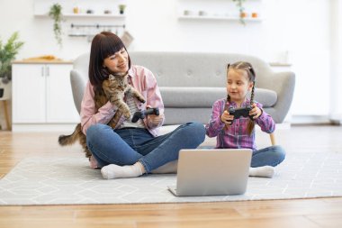 Samimi bir aile bağı, rahatlama ve kapalı mekan aktivitesi anı. Anne ve kızı dizüstü bilgisayarla yerde oyun oynayarak eğleniyorlar. Kedi, konforlu ev ayarlarına eğlenceli öğeler ekler.