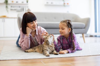 Anne ve kızı kedileriyle sıcak oturma odasında kaliteli zaman geçiriyorlar. Yerde kedi besleyip sıcak bir aile ortamı yaratıyorlar. Arka planda modern mobilya.