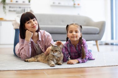 Anne ve kızı evcil kedileriyle oturma odasında yerde yatıyorlar. Aile kaliteli zaman geçiriyor, sıcak ev ortamında sevgi ve arkadaşlığın altını çiziyor.