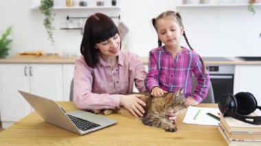 Dizüstü bilgisayarda çalışan bir kadın. Çocuk evcil hayvanla iyi vakit geçiriyor. Anne ve kızı mutfakta kedi beslerken gülümsüyor. Aile bağları olan rahat bir ev ortamı.