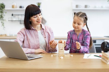 Anne ve kızı evde Jenga oynayarak güzel vakit geçiriyorlar. Gülümseyen kadın ve mutlu çocuk oyundan zevk alıyor ve rahat mutfak ortamında dizüstü bilgisayarla bağ kuruyor ve malzeme çiziyor..