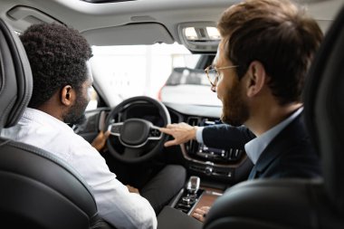 Alıcı direksiyonu tutarken, satıcı da aracın detaylarına dikkat çekerek profesyonel hizmet sunuyor. Beyaz araba satıcısı Afrika kökenli Amerikalı alıcıyla birlikte aracın içinde oturup özellikleri tartışıyor..