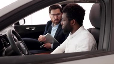 Mutlu müşteri yeni araba alımını galerideki profesyonel satıcıdan öğreniyor. Kafkasyalı araba satıcısı içerideki Afrikalı Amerikalı alıcıya araç özelliklerini açıkladı.