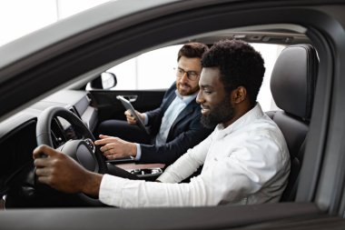 Mutlu müşteri yeni araba alımını galerideki profesyonel satıcıdan öğreniyor. Kafkasyalı araba satıcısı içerideki Afrikalı Amerikalı alıcıya araç özelliklerini açıkladı.