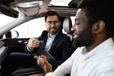 Profesyonellik, güven ve memnuniyet ile karakterize edilen etkileşim. Beyaz araba satıcısı Afrika kökenli Amerikalı alıcıyla birlikte araçta oturuyor, araba anahtarlarını teslim ediyor, başarılı bir satın almayı temsil ediyor..