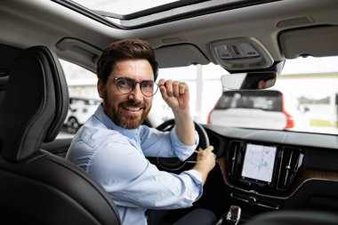 Gülümseyen iş adamı galeride arabada oturuyor. Gözlük takan, iş kıyafetleri giyen ve direksiyon tutan bir adam. Arkaplanda araçlar olan parlak bir iç mekan.