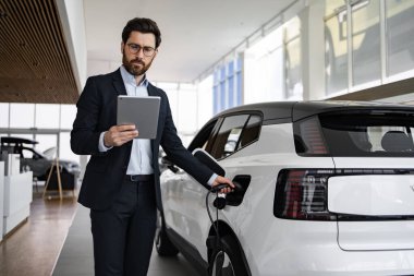 Takım elbiseli satıcı, tablet kullanarak galeride elektrikli araba satıyor. Sürdürülebilir ulaşım ve modern teknolojiyi tanıtırken elektrikli araç şarj ediliyor.