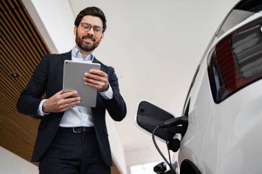 Modern araç galerisinde elektrikli araba satmak için dijital tablet kullanan bir satıcı. İş alışverişi sırasında çevre dostu teknoloji sergileyen profesyonel takım elbiseli..