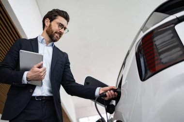 Profesyonel araba satıcısı modern galeride elektrikli araba sergiliyor, tablet tutuyor ve müşteriyle takılıyor. Çevre dostu araçlar, otomotiv teknolojisi ve sürdürülebilir ulaşım kavramı.