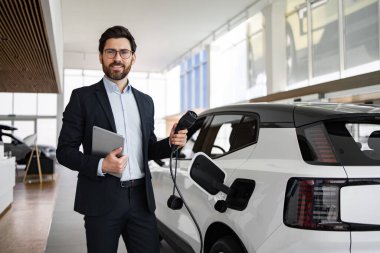 Bir satıcının elinde tablet ve elektrikli araba şarj prizi bulunan bir galeride satıcı, çevre dostu araçlar ve yeni araba teknolojisi sergiliyor. Arka planda çeşitli elektrikli arabalarla modern sergi salonu.