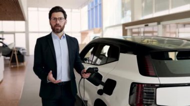 Gülümseyen galerici modern araba bayiliğinde elektrikli araba sergiliyor. Araba satışı için tablet kullanan profesyonel bir satıcı. Çevre dostu teknolojiyi vurguluyor.