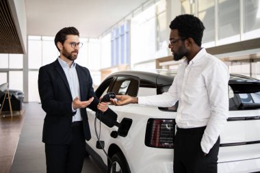 Satıcı, araba galerisindeki iş adamlarına elektrikli cip satıyor. Ticari işlemler ve sürdürülebilir araç yatırım kavramı. Elektrikli arabaların özellikleri ve faydaları hakkında konuşan erkekler.