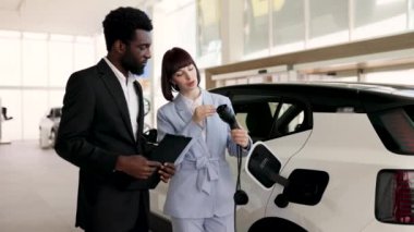 Gülümseyen araba satıcısı araba galerisindeki iş kadınına elektrikli cip satıyor. Satıcı araç özelliklerini açıklarken iş kadını elinde elektrikli şarj aleti tutuyor. Modern araba, çevre dostu ulaşım..