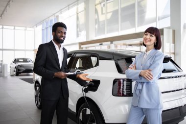 Araba galerisindeki iş kadınına elektrikli SUV gösteren bir satıcı özellikleri ve özellikleri tartışıyor. Parlak sergi salonu, beyaz modern elektrikli arabalar, gösterişli iç mekanlar, iş kıyafetleri, gülen yüzler..