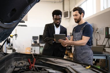 Tamirci, atölyede dijital tablet kullanarak iş adamıyla araba tamir detaylarını tartışıyor. İşbirliği, teknoloji ve otomobil hizmeti kavramı.