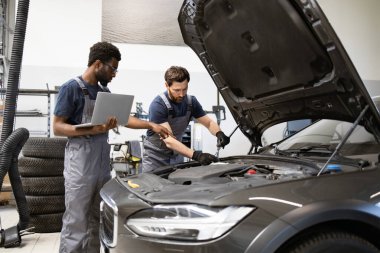 Atölyedeki tamirciler, teşhis için dizüstü bilgisayar kullanarak araç motoru tamir ediyorlar. Ekip çalışması ve teknoloji otomotiv tamir servisinde. Modern araba bakımı ve denetimi.