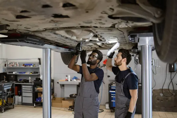 stock image Mechanics performing car maintenance under lift in professional auto repair shop, collaborating on vehicle repair, wearing work uniforms, and using tools for inspection and fixing.