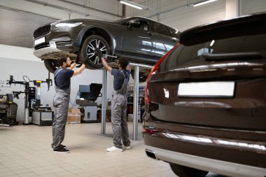 Modern oto tamirhanesinde profesyonel tamirciler araba tamir ediyor. İki teknisyen asansördeki aracı inceliyor. Temiz atölye ortamında otomotiv hizmeti, bakım ve takım çalışması.