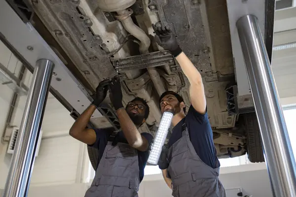 stock image Two auto mechanics wearing uniforms examining car underside using tools and light. Professional technicians performing vehicle maintenance and repair service in modern garage workshop.