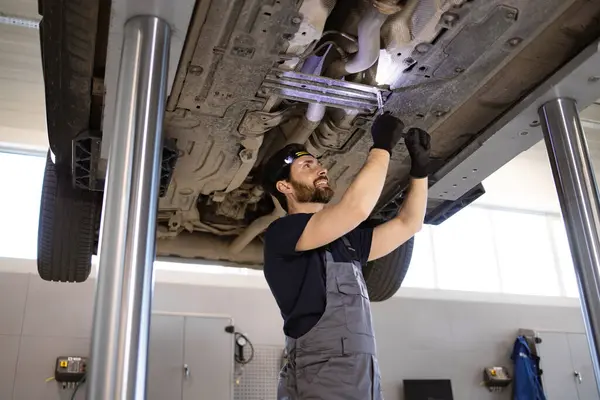 stock image Skilled auto mechanic repairing car undercarriage in service garage. Wearing gloves, professional working with tools. Car raised on lift, ensuring safety and precision.