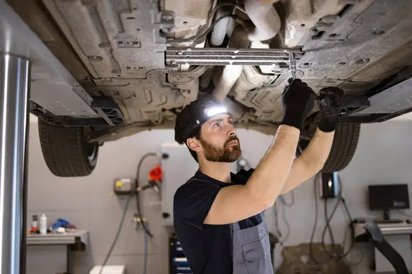 stock image Dedicated mechanic performing maintenance on car in professional garage. Wearing headlamp and gloves, focusing on vehicles undercarriage for repair work.