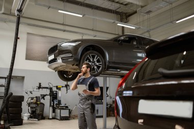 Atölye üniformalı tamirci modern oto tamirhanesinde dijital tablet kullanarak kaldırılmış arabayı inceliyor. Profesyonel otomotiv bakımı ve teşhisi. Alet ve ekipmanlarla dolu bir atölye.